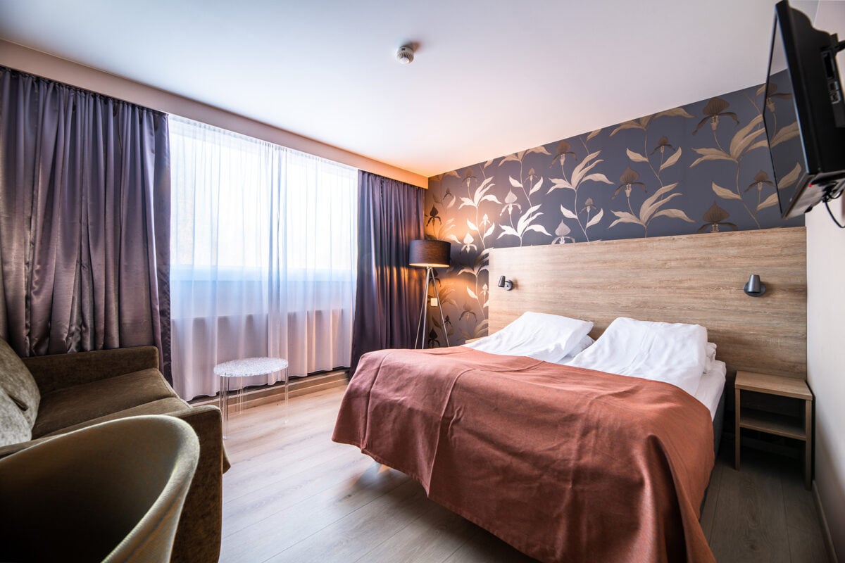 Moderne hotellrom med dobbeltseng, brunt teppe og stilfullt tapet, belysning fra gulvlampe ved gardiner.