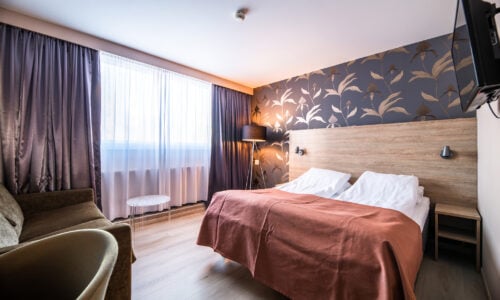 Moderne hotellrom med dobbeltseng, brunt teppe og stilfullt tapet, belysning fra gulvlampe ved gardiner.