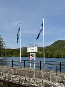 Veiskilt ved vannkanten i Lunde med livbøye. To flaggstenger ved siden av. Skog i bakgrunnen. Klar blå himmel.