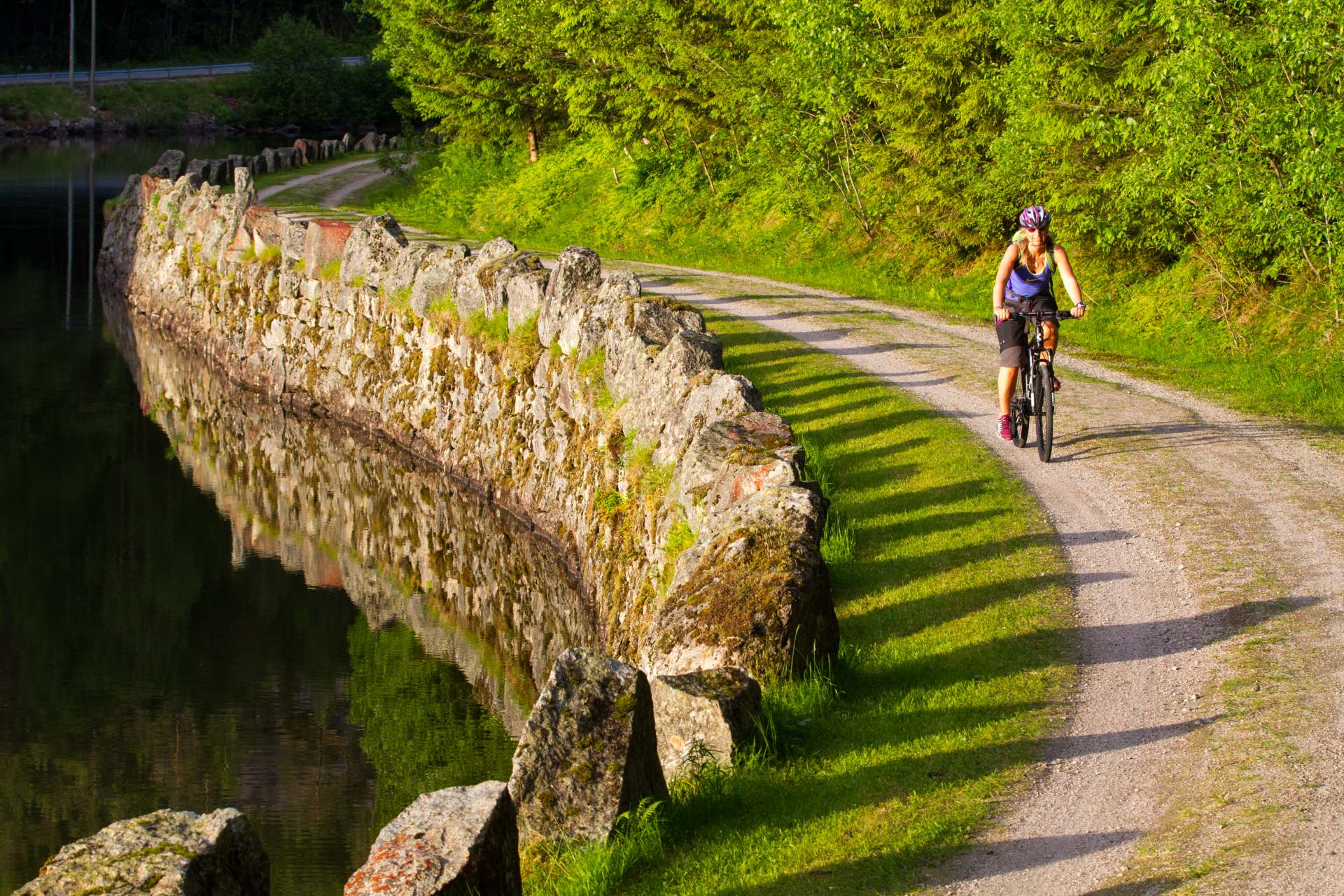 Sykklist på grusvei ved steingjerde, omgitt av grønn skog og vannspeiling i solskinn.