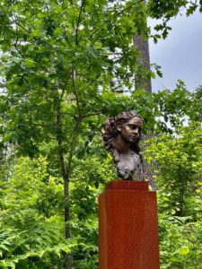 Byste av kvinne på sokkel i skoggløtt, omringet av grønne trær.