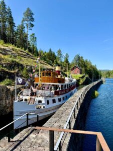 Båt ved innsjø omgitt av skog og hus. Klar blå dag. Turister på dekk. Skandinavisk natur.