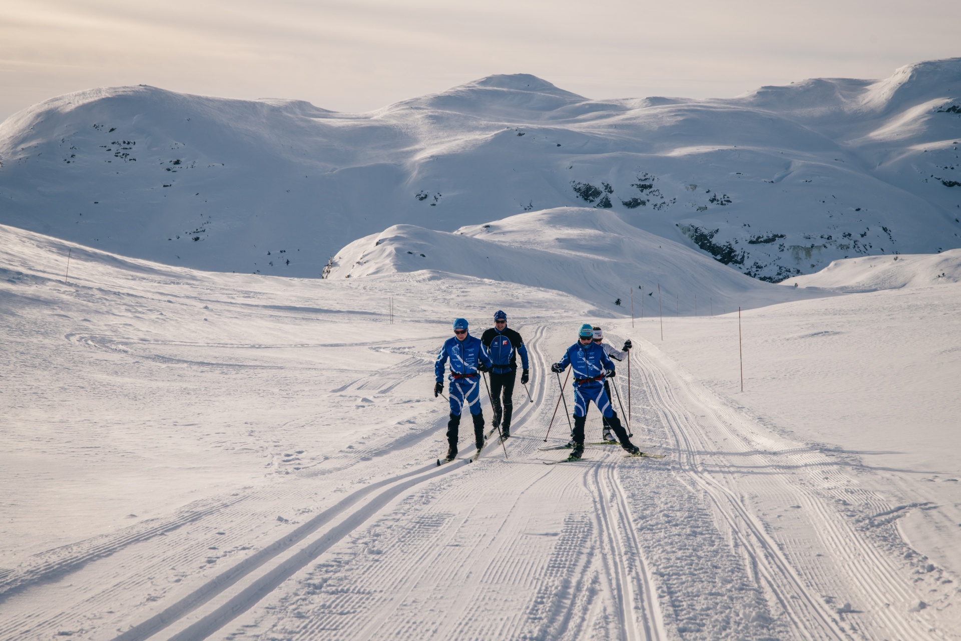 Skiløpere i blått konkurrerer i langrenn på snødekkede fjell med vakker himmelbakgrunn.