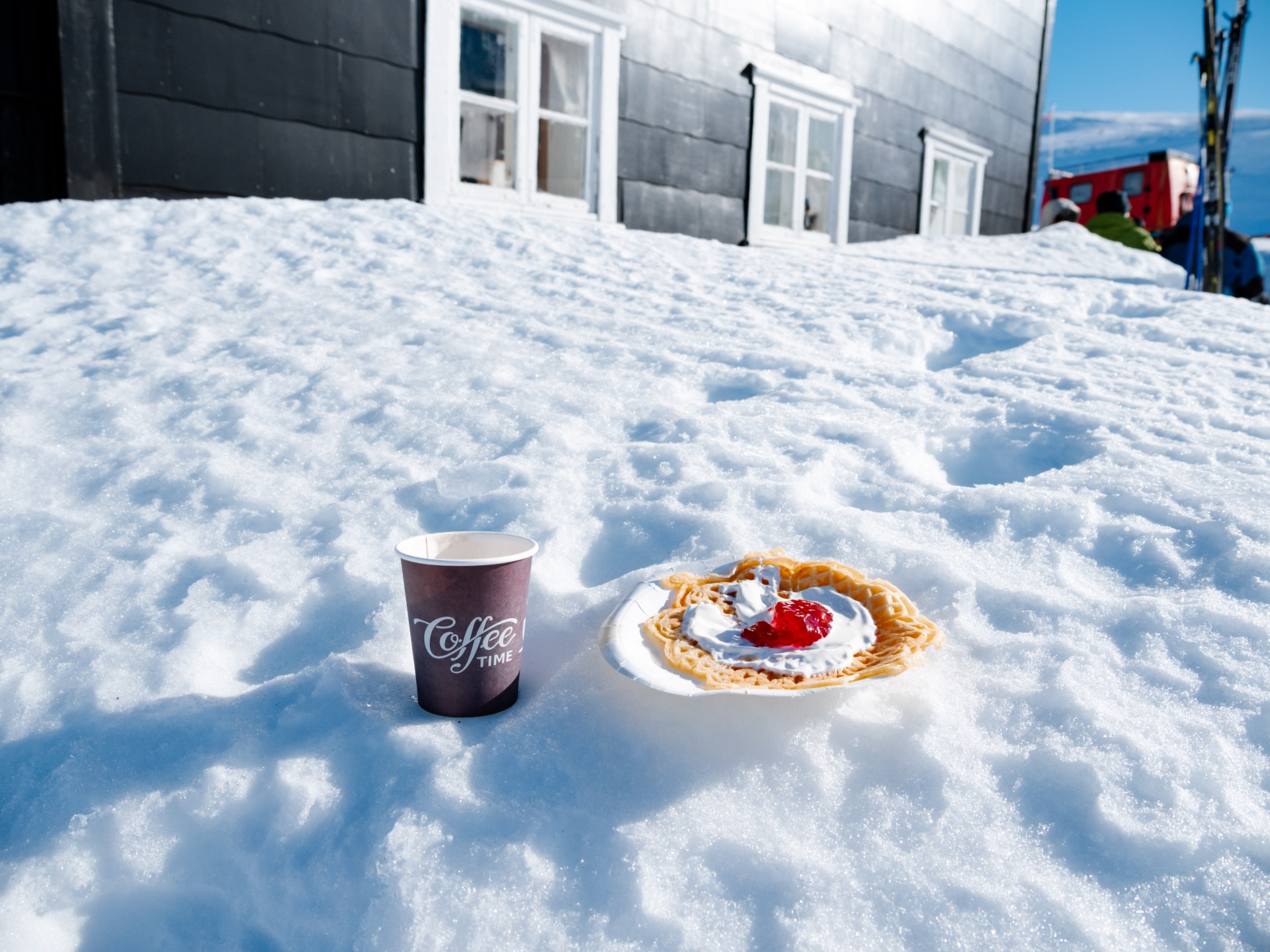 Vaffel med syltetøy og krem på snøen ved siden av en kaffekopp, foran et svart hus.