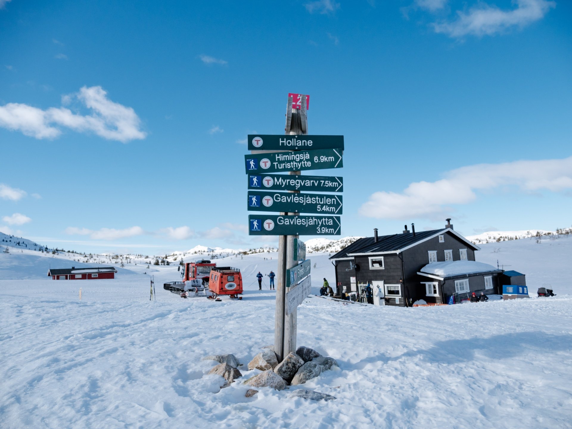 Skilt med snødekte turstier i fjellet, hytter og weasel i bakgrunnen under en klar blå himmel.