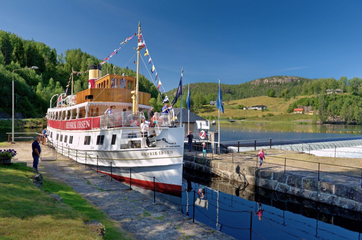 Bilde av båten Henrik Ibsen ved en havn, med besøkende og vakker natur i bakgrunnen. Flott sommerdag.