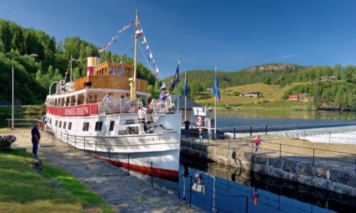 Bilde av båten Henrik Ibsen ved en havn, med besøkende og vakker natur i bakgrunnen. Flott sommerdag.