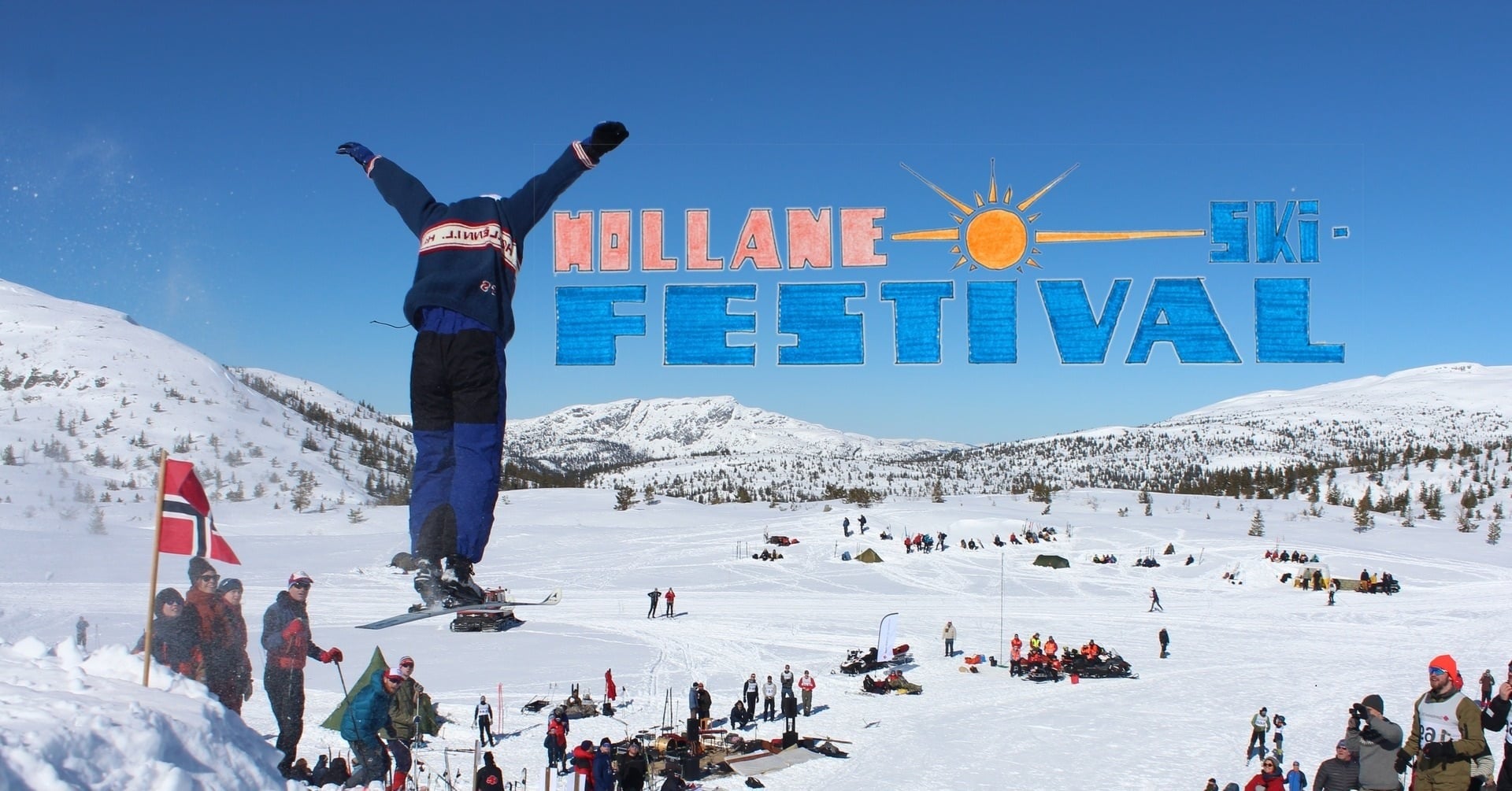 En skikjører hopper under Hollane Ski-Festival, med snødekte fjell og tilskuere i bakgrunnen.
