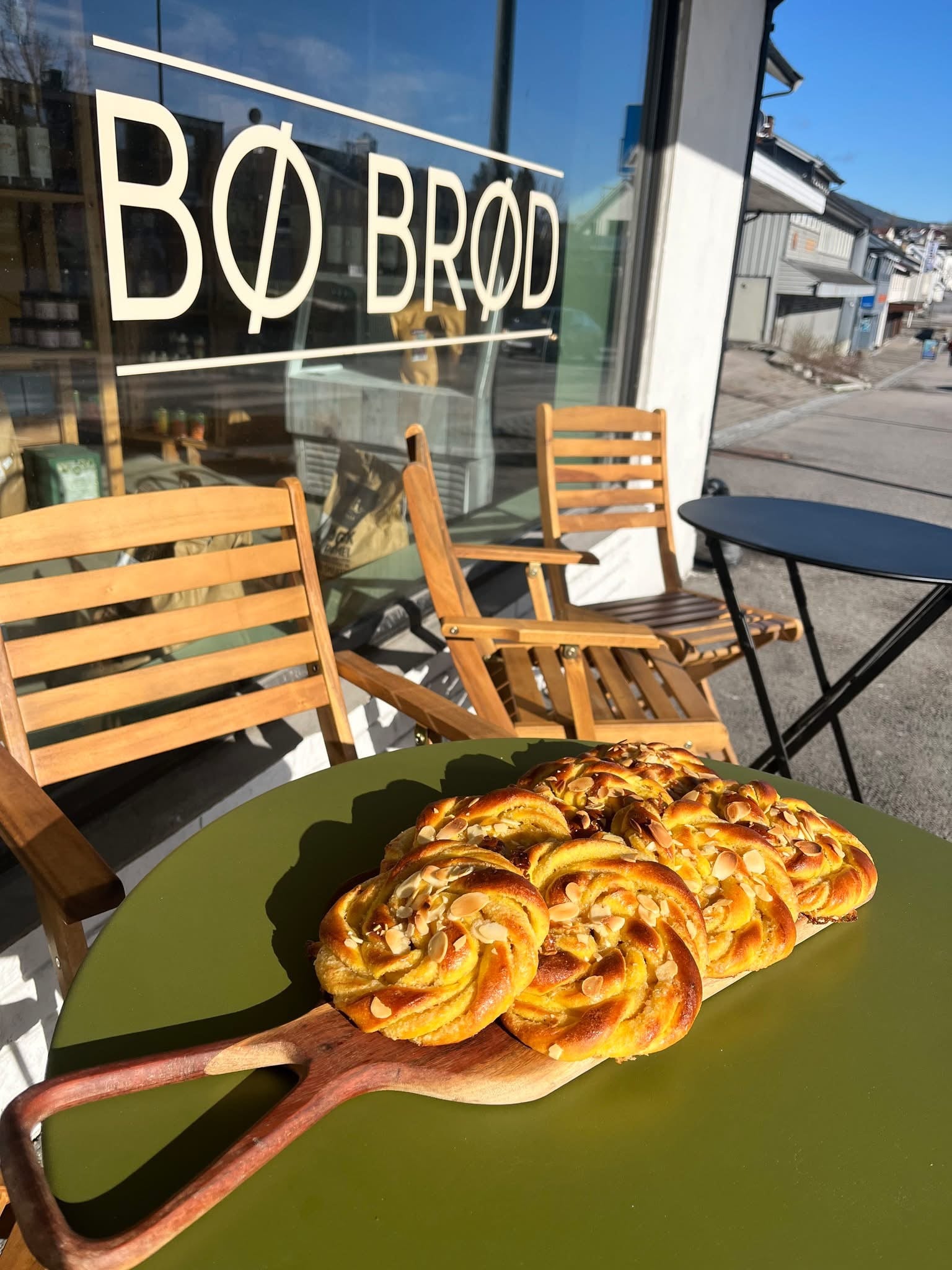 Delikat kanelbolle på en trefjøl, foran BØ BRØD-butikken, med solfylt uteområde og koselige stoler.