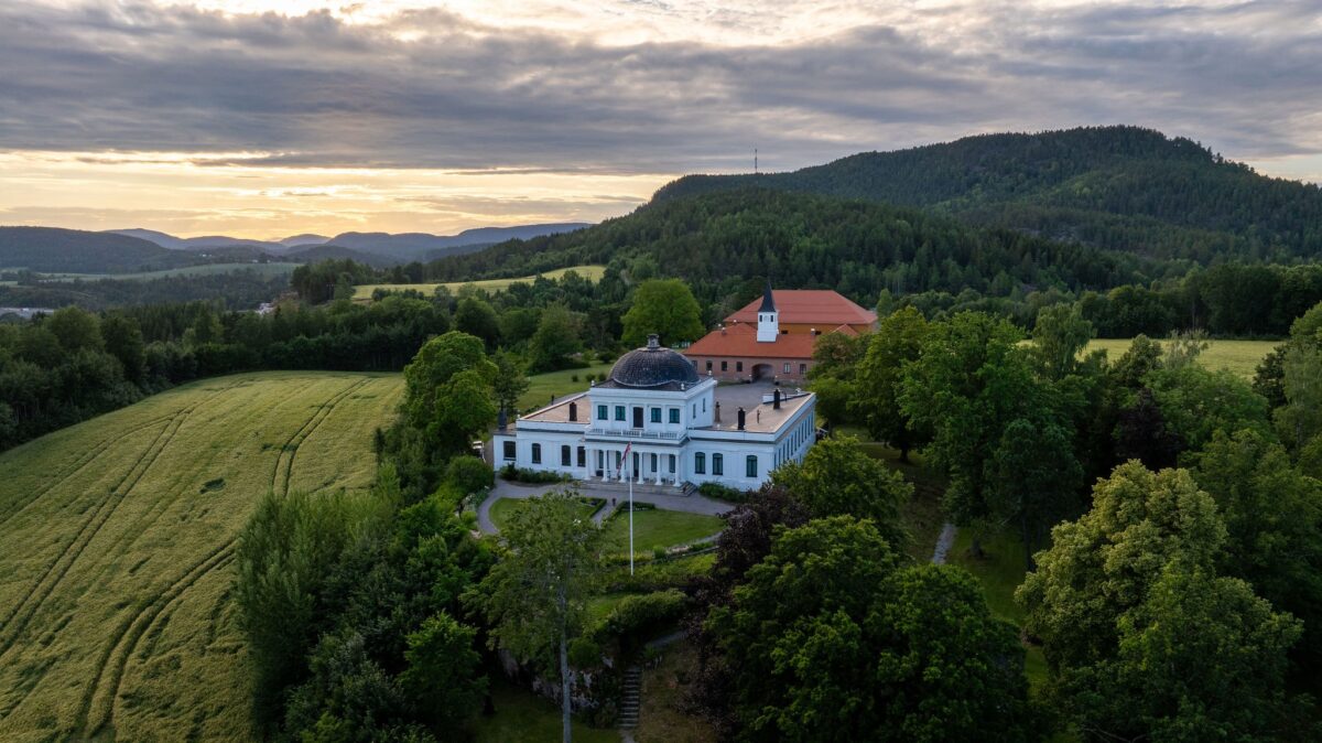 Luftbilde av en vakker bygning omgitt av grønne åser og trær, med dramatisk himmel ved solnedgang.