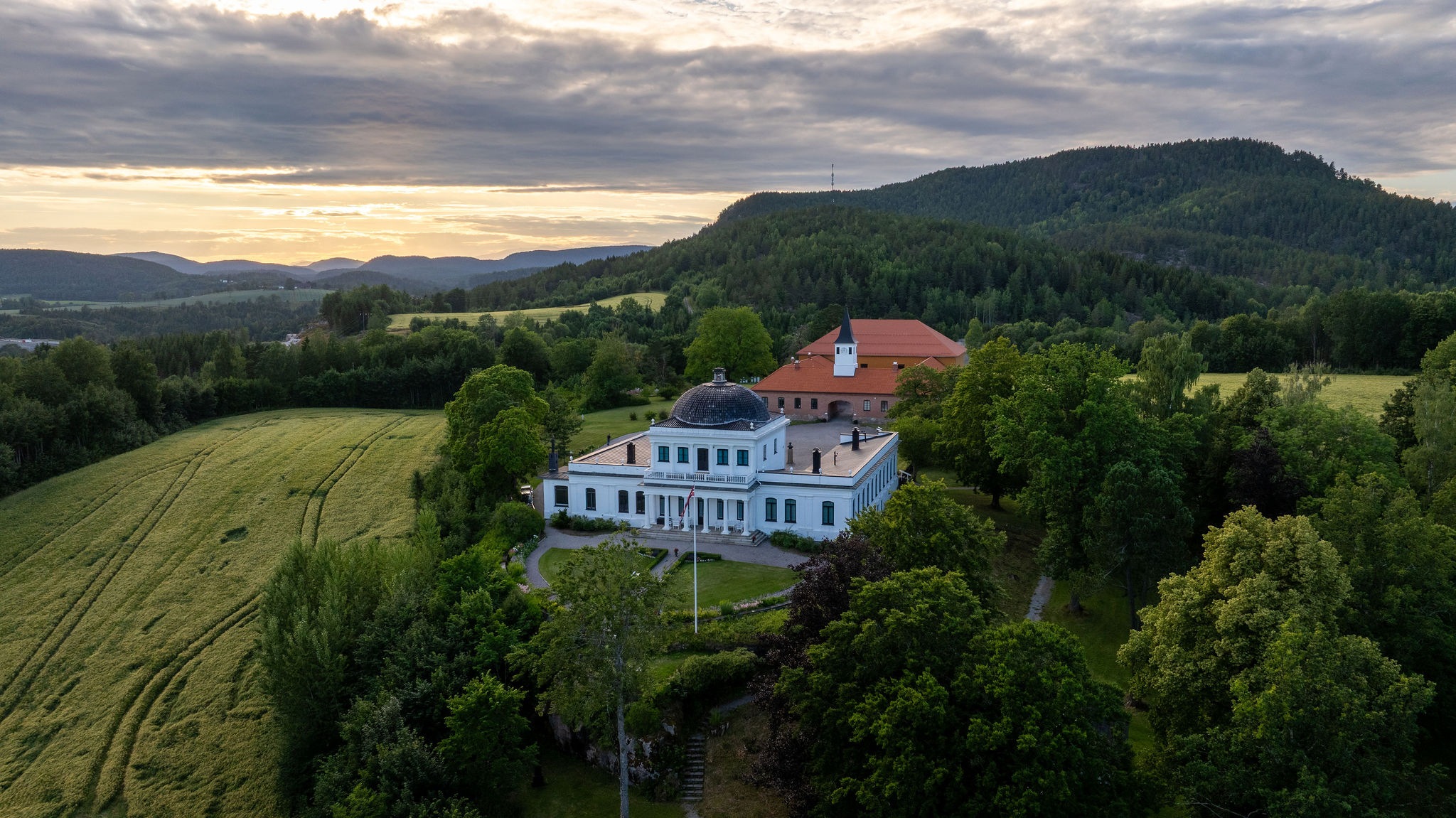 Luftbilde av en vakker bygning omgitt av grønne åser og trær, med dramatisk himmel ved solnedgang.