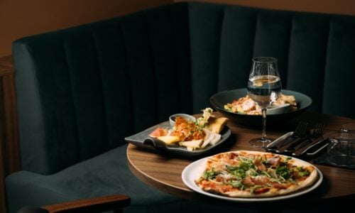 Tallerken med pizza, ost og salat på bord i restaurant, med vinglass og bestikk, moderne mørk interiør.