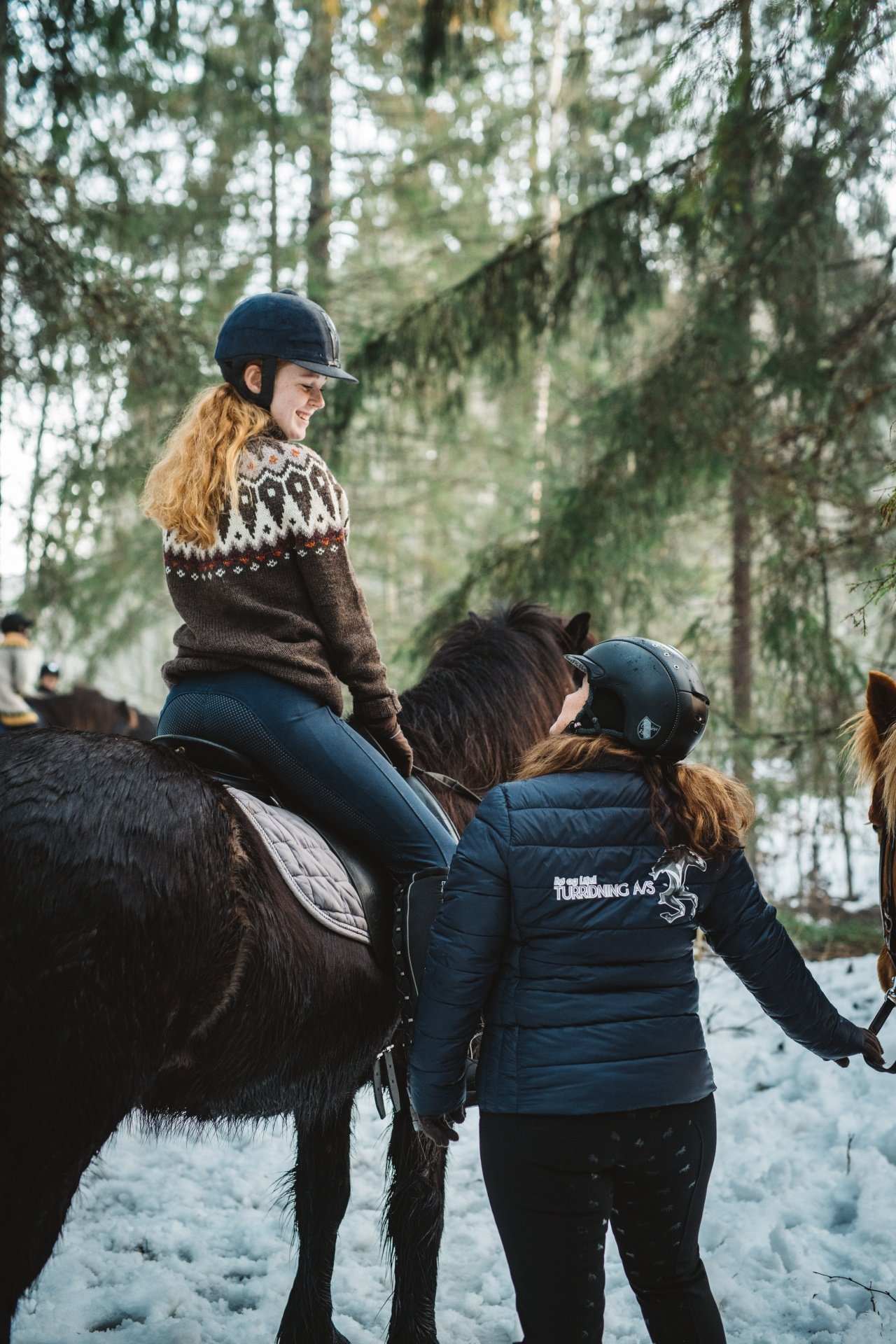 Kvinne rir hest i vinterlandskap med instruktør, iført ullgenser og hjelm i skogen.