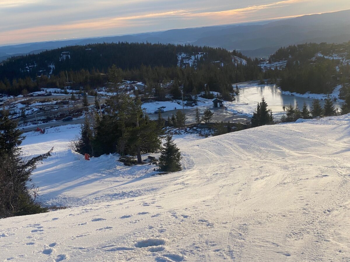 Snødekt landskap med fjell og innsjø i horisonten, omgitt av skog under soloppgang. Perfekt for vinteraktiviteter.