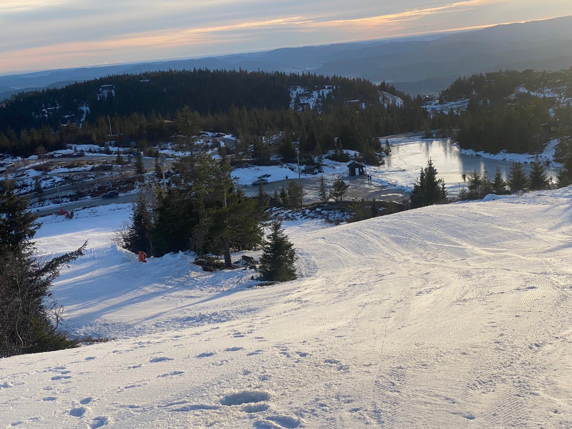 Snødekt landskap med fjell og innsjø i horisonten, omgitt av skog under soloppgang. Perfekt for vinteraktiviteter.