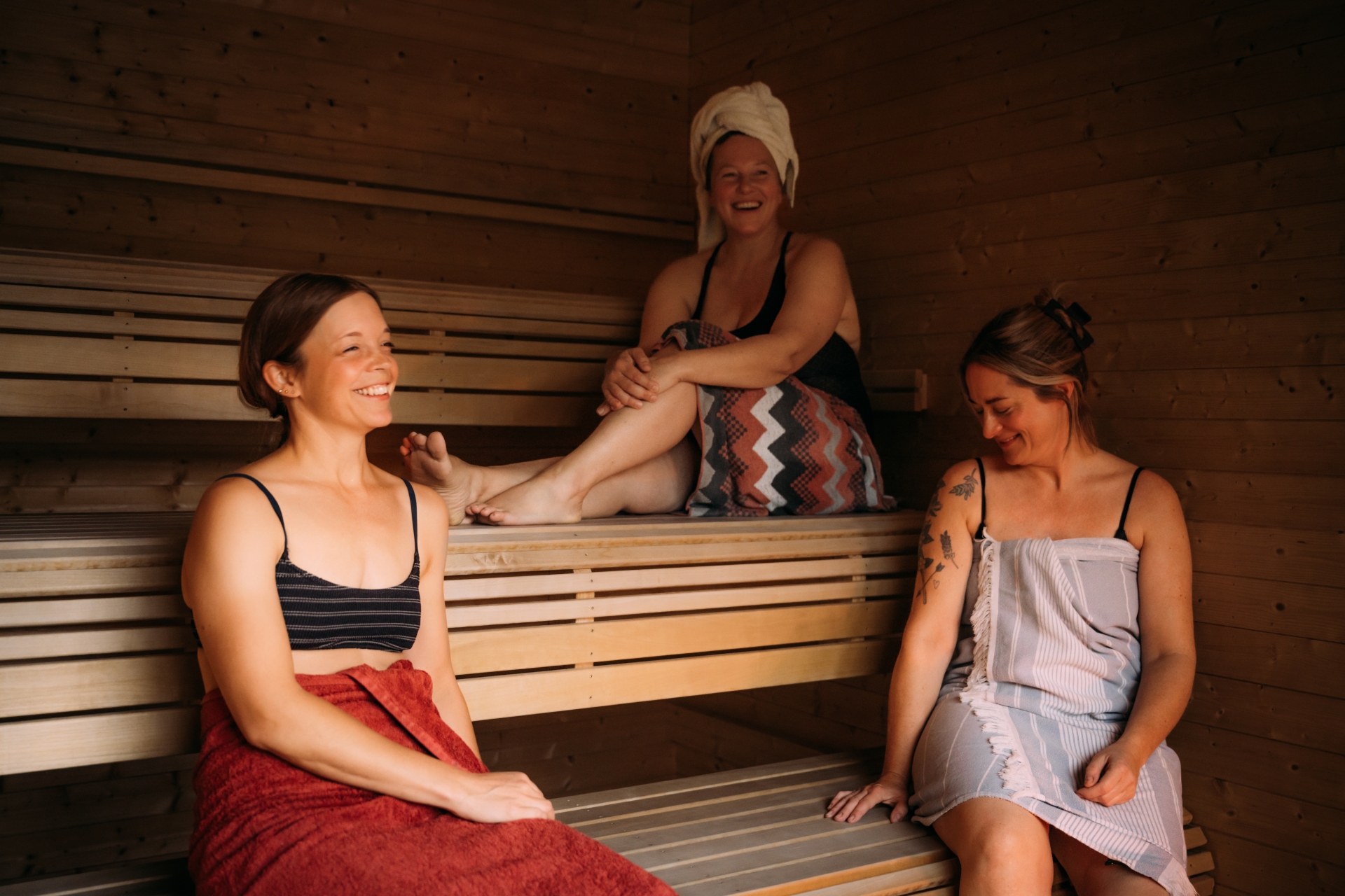 Tre kvinner nyter avslappende tid i en badstue, omringet av trevegger, iført badetøy og håndklær.