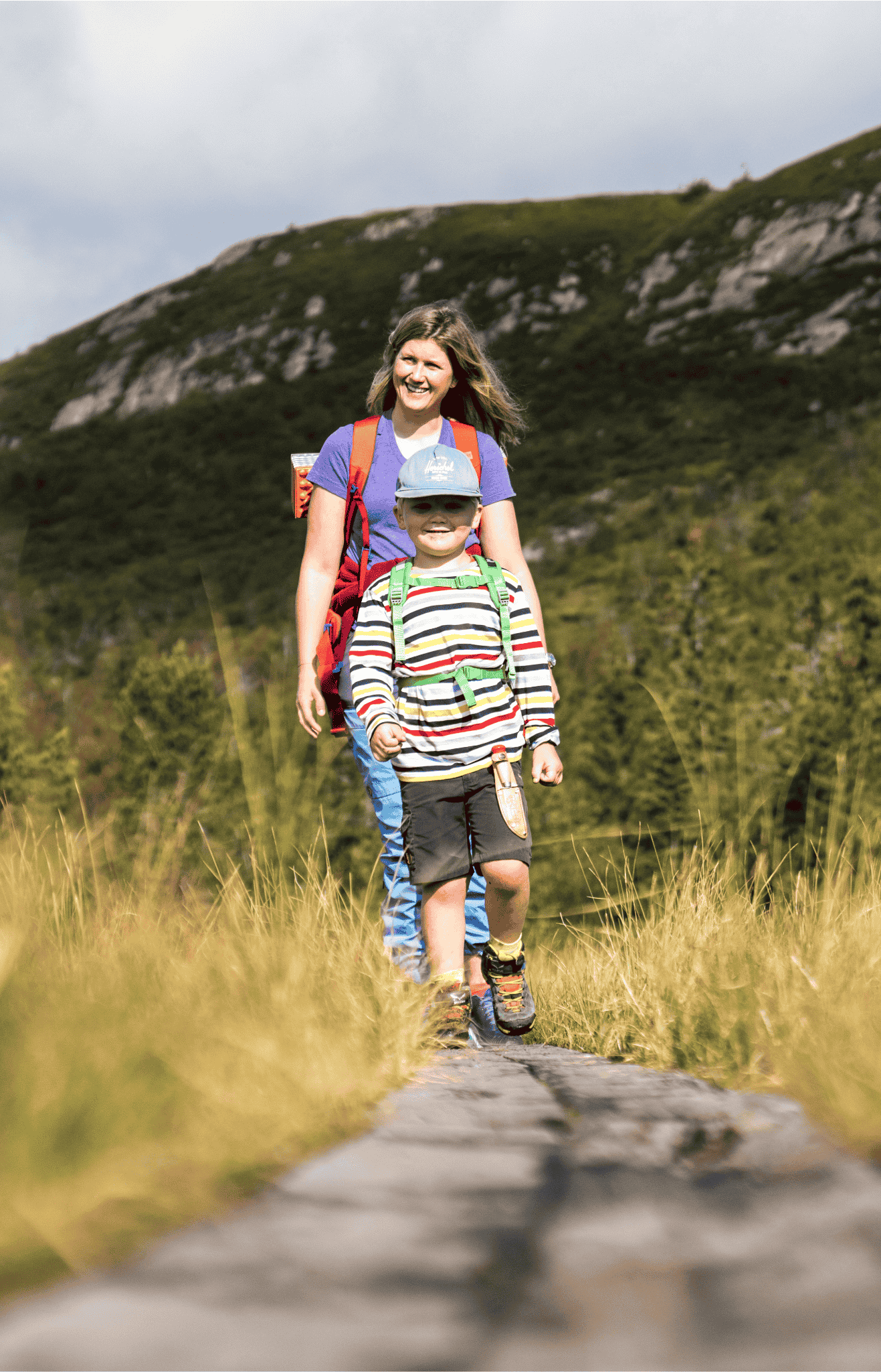 Mor og barn går sammen på tur i fjellet, omgitt av grønn natur og sollys. Perfekt for familieturer!