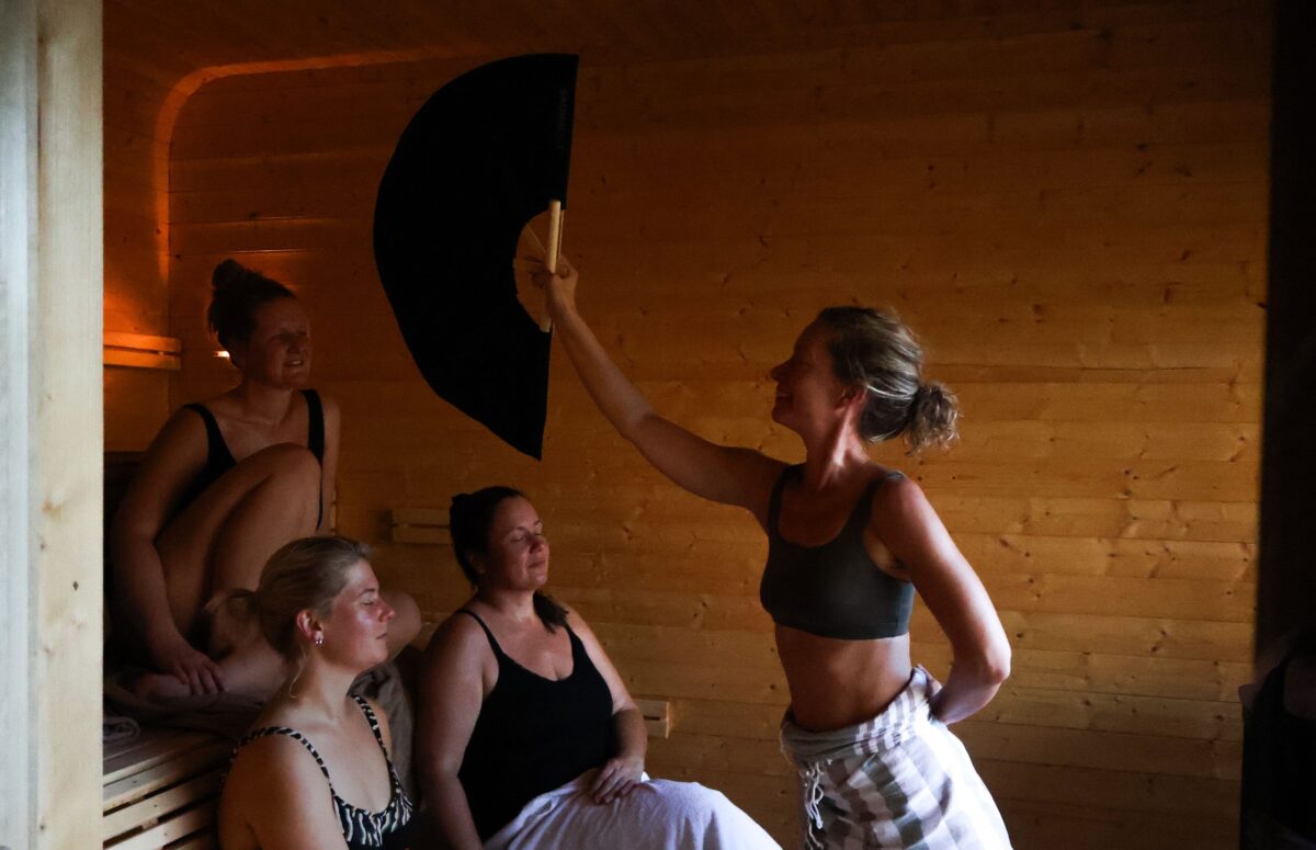 Kvinner i badstue som nyter avkoppling, en av dem holder en stor vifte for å sirkulere varmen.
