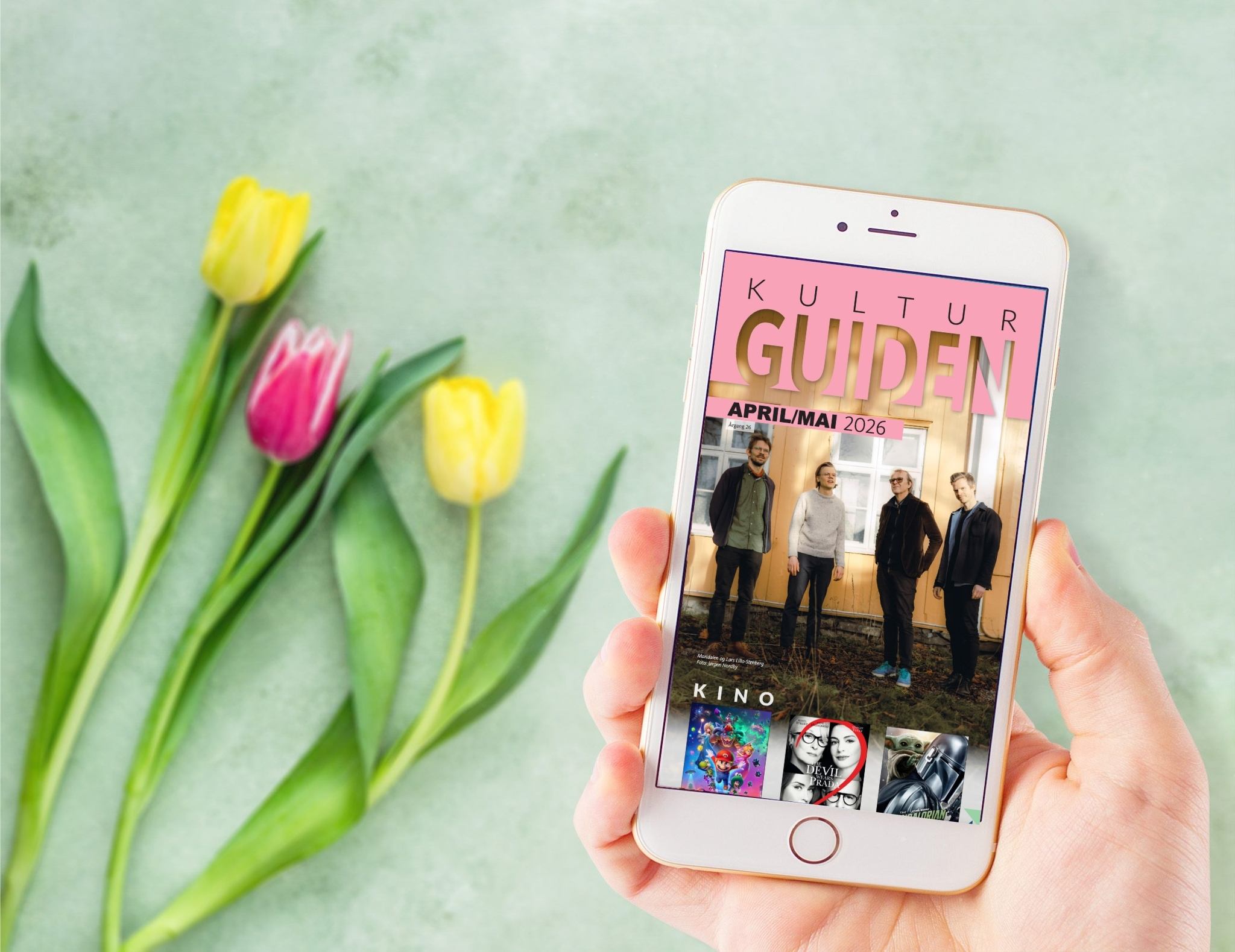 Hånd som holder smarttelefon med Kulturguiden for april/mai 2026 og tulipaner i bakgrunnen.