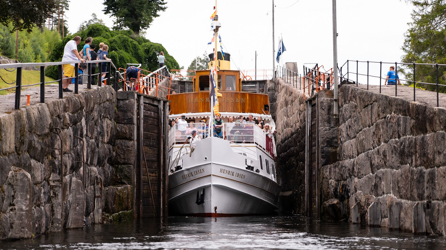 Båten Henrik Ibsen passerer en sluse med mennesker som ser på fra siden. Vakkert landskap omgir scenen.