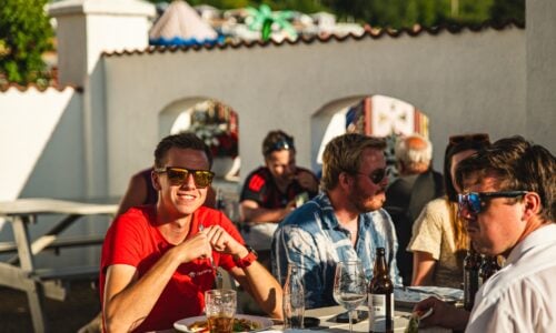 Menn og kvinner sitter ute og nyter mat og drikke på en solfylt sommerdag. Stemning av glede og samvær.
