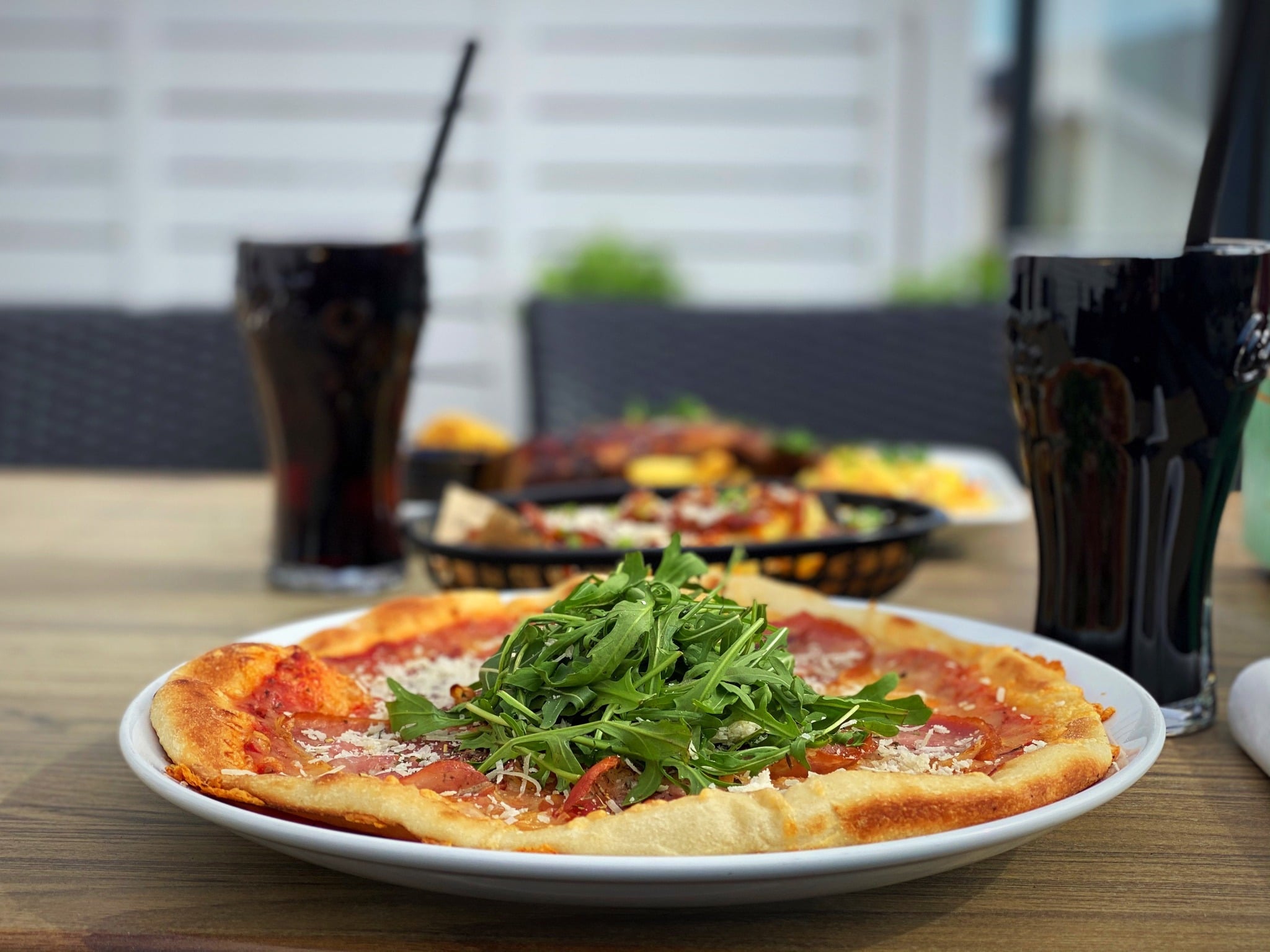 Lekker pizza med ruccola på tallerkenen, med brus i bakgrunnen på en koselig terrasse.