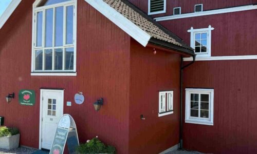 Rødt hus med store vinduer, en gårdsskilt foran og blomster i krukker. Vakker norsk arkitektur under blå himmel.