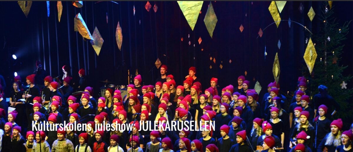 Kulturfaglig juleshow med barn som synger, iført røde luer, på scenen i et festlig miljø.